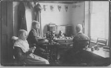 SA0006 - Three women sewing; Emma Neale is in the center. Photo shows a sewing machine and sewing paraphernalia. Caption on the back.